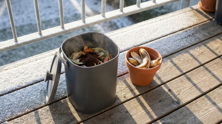 A sunlit apartment balcony showcases a compact, sleek bokashi composting bin in muted charcoal gray.
